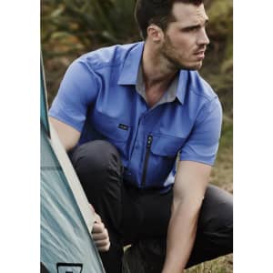 A short-sleeve, blue button-up shirt with one chest pocket and logo, worn by a man outdoors near a tent.