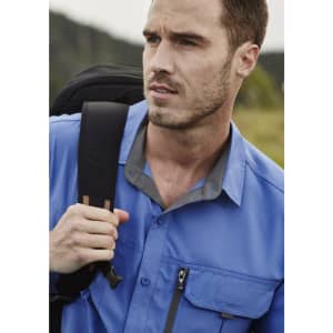 A man wearing a blue short sleeve button-up outdoor shirt, carrying a backpack, with a scenic background.