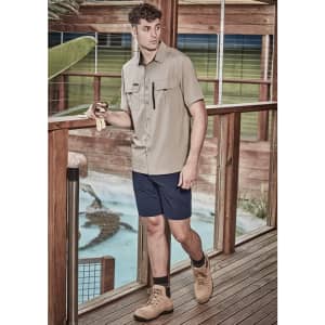 A man wearing a light brown short sleeve button-up shirt and navy shorts, standing on a wooden deck.