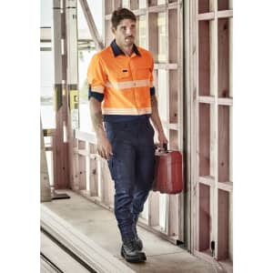 A man wearing an orange long sleeve segmented tape work shirt and navy pants, holding a red toolbox in a construction setting.