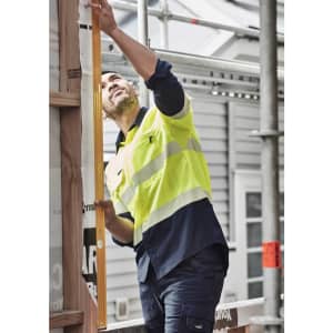 A man wearing a hi-vis yellow and navy long sleeve work shirt examines a construction site using a level tool.