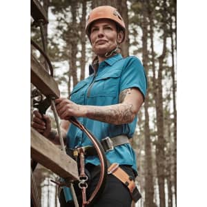 A woman wearing a blue short sleeve outdoor shirt with a harness, set against a forest background.