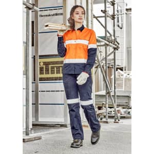 A woman in an orange and navy long sleeve industrial shirt, holding timber, wears work gloves and sturdy boots.