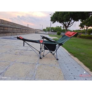 A green and black camping lounge chair with red accents, set on stone paving near grass and trees.