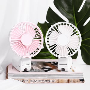 A pink and white rechargeable folding hand-held fan placed on top of a book, against a backdrop of green leaves.