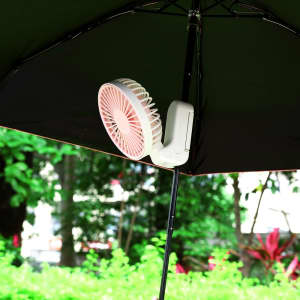 A rechargeable, folding hand-held fan in white and pink, attached to a dark umbrella with greenery in the background.
