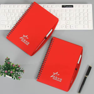 A red spiral notebook with a pen, featuring a logo, placed on a desk with a keyboard and a small plant.