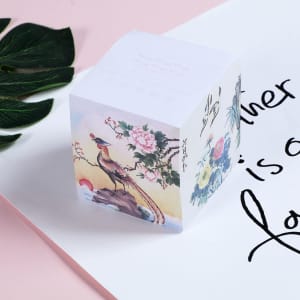 A square sticky note cube featuring floral and bird designs in pastel colours, placed on a pink surface.