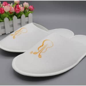 A pair of white disposable hotel slippers with an embroidered design, placed on a grey surface near decorative flowers.