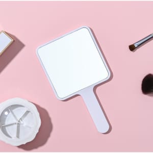 A square handheld mirror with a white handle, placed on a pink background alongside beauty tools.