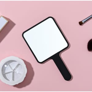 A square handheld mirror with a black handle, placed on a pink surface alongside beauty tools.