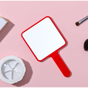 A square handheld mirror with a red frame, placed on a pink surface alongside cosmetic items.
