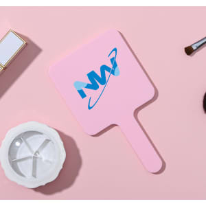A square handheld mirror in pink with a branded logo, placed on a light pink surface alongside makeup accessories.