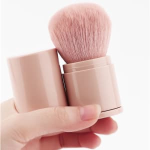 A retractable makeup brush in a pink casing, featuring soft bristles, held in a hand.