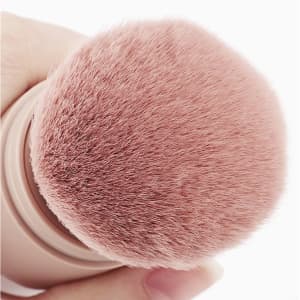 A retractable makeup brush with a soft pink brush head, featuring a sleek holder in neutral tones and a logo.
