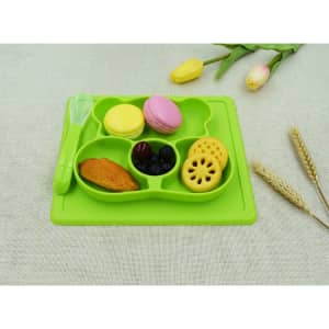 A green non-slip children's plate with compartments holding various treats, including macarons, fruit, and a cookie.