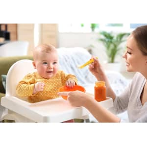 A baby sits in a high chair, smiling at a woman holding a yellow spoon and a jar of orange food.
