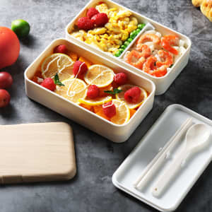 A double-layer lunch box set in white containing fruits, vegetables, scrambled eggs, and seafood, with a wooden lid and utensils.