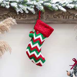 A Christmas stocking with a red top and a green, white, and red zigzag pattern, featuring a pom-pom.