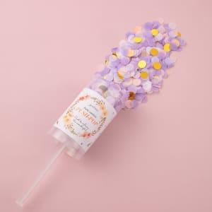 A clear push pop filled with purple, pink, and gold confetti against a pink background, featuring a decorative label and a straw.
