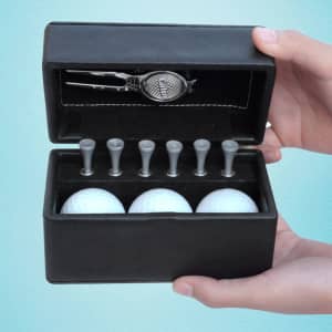 A black golf set box containing three white golf balls, six wooden tees, and a silver divot tool.