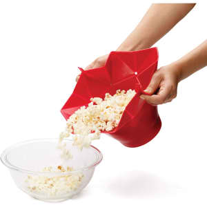 A red popcorn bucket spilling popcorn into a glass bowl, featuring a geometric design and held by a hand.