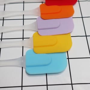 A set of cream spatulas in red, orange, purple, yellow, and blue, with transparent handles, arranged in a row.