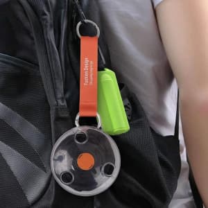 A folding telescopic storage bag in black and transparent with an orange strap, attached to a backpack alongside a green item.