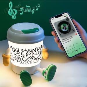 A coffee cup-shaped lamp Bluetooth speaker with a smiling face, green details, and a smartphone playing music nearby.