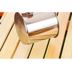 A stainless steel camping mug with a folding handle, resting on wooden planks. The mug has a shiny finish and a logo.