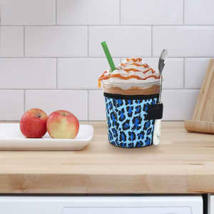 A blue leopard print ice cream cup holder with a spoon, featuring a drink and two apples on a wooden countertop.