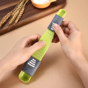 An adjustable measuring spoon in green and grey, with measurement markings, held in two hands against a light background.