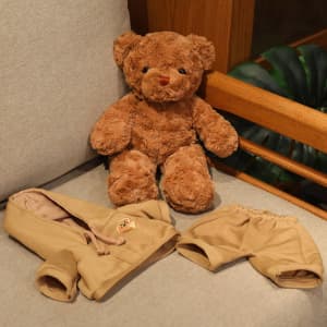 A brown plush teddy bear accompanied by a tan outfit consisting of a hoodie and shorts, placed on a light-coloured surface.