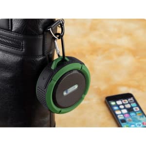 A green waterproof Bluetooth speaker hangs from a black bag, with a smartphone resting nearby. The speaker has a logo.