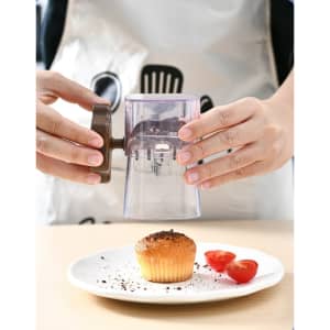 A manual mashing and grinder in clear plastic with a brown handle, used above a plate with a cupcake and tomatoes.