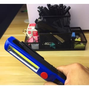A handheld work light in blue with a yellow LED strip, positioned in front of a pen holder filled with various items.