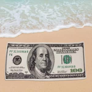 A beach towel featuring a large printed image of a hundred-dollar bill, set on a sandy beach with water.