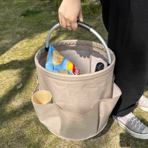 A portable tool basket in beige, featuring pockets and a handle, holding snacks and a drink cup.