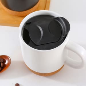 A ceramic mug with a cork base, featuring a black lid, filled with coffee beans on a wooden surface.