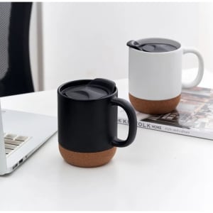 A ceramic mug with a cork base in black and white, featuring a lid, placed beside a laptop on a table.