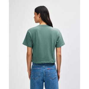 A green women's boxy t-shirt, viewed from the back, paired with light-coloured jeans. It features a branding logo.