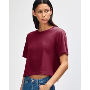 A woman with curly hair wearing a burgundy boxy t-shirt, styled with light blue jeans. The shirt has a logo.