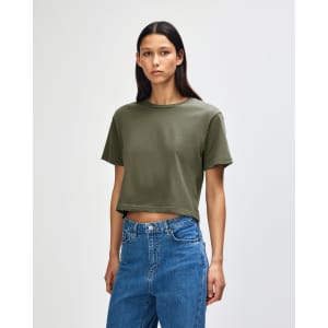 A woman wears an olive green boxy t-shirt, styled with light blue jeans, against a plain background.