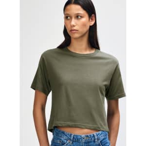 A woman in an olive green boxy T-shirt, styled with high-waisted jeans and curly hair. The shirt has a logo.