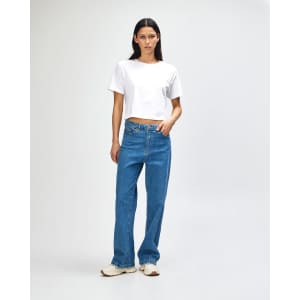 A woman stands wearing a white boxy t-shirt and light blue wide-leg jeans, featuring a logo on the attire.