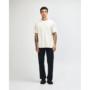 A unisex heavyweight cotton T-shirt in cream, paired with blue cargo pants and sneakers. The model has curly hair and a neutral pose.