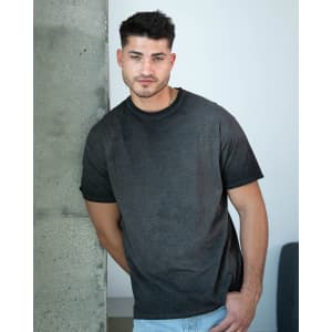 A man wearing a dark grey Colortone® Oil Wash T-Shirt, leaning against a concrete wall. The shirt has a logo.