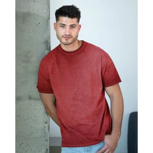 A man posing by a concrete wall, wearing a red Colortone® Oil Wash T-Shirt. The shirt has a casual style and relaxed fit.