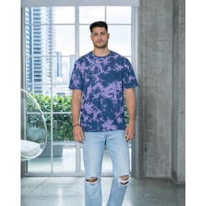 A man stands in a modern space wearing a purple and navy tie-dye T-shirt and ripped jeans. The shirt features a logo.