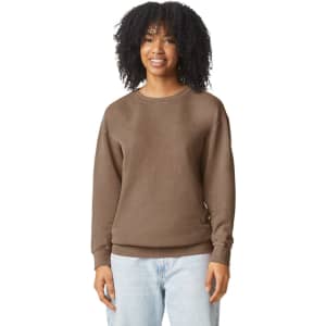 A brown crewneck sweatshirt featuring a relaxed fit. The model has curly hair and is smiling. The sweatshirt has a logo.